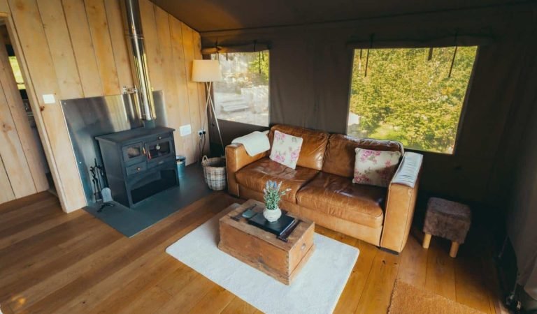 Spacious glamping living room with log burner, tan leather sofa and large windows overlooking woodland.